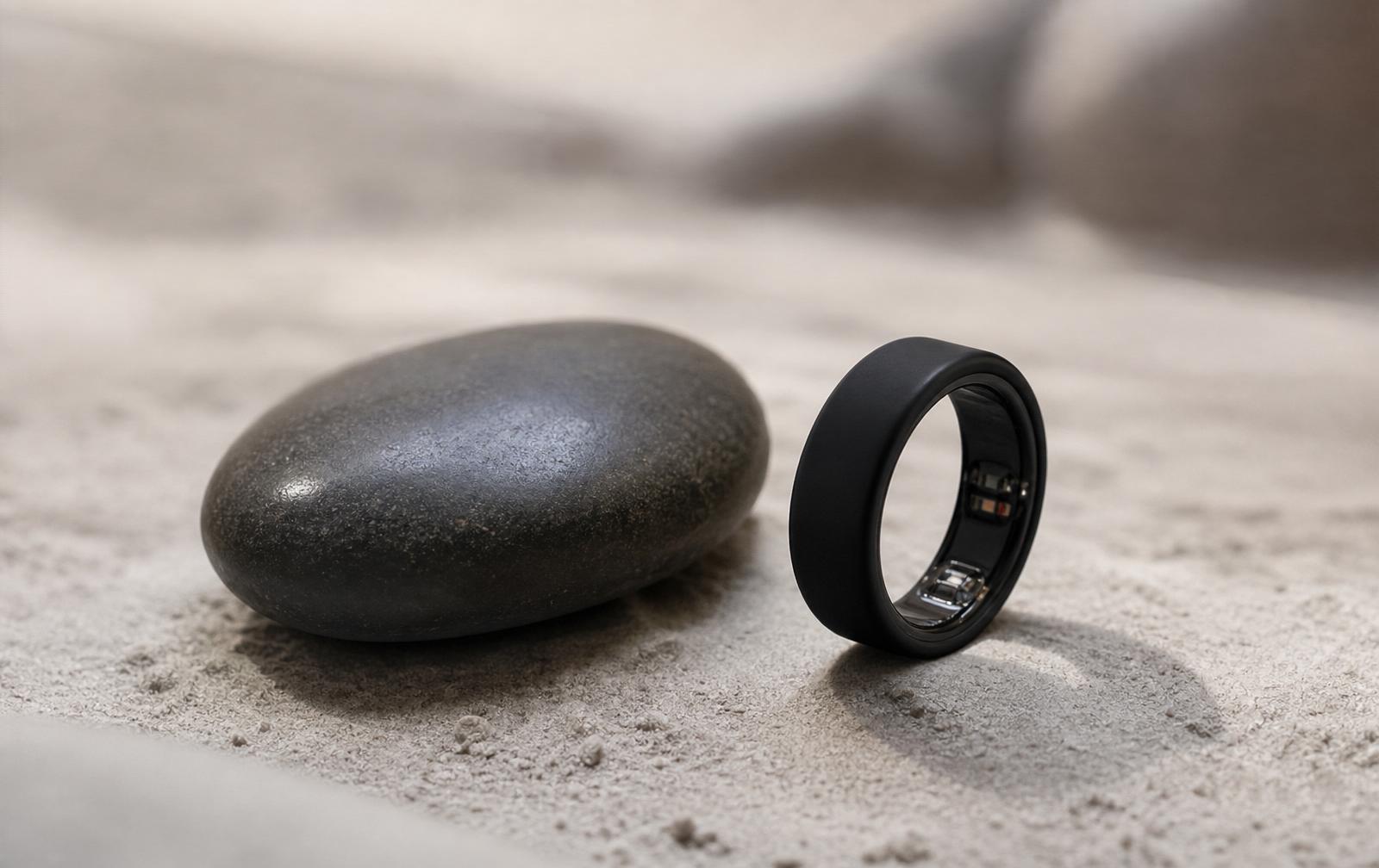 A smooth dark polished river stone beside a sleek minimal biometric ring on clean textured light gray soil